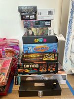 Stack of board games, puzzles, and electronic games showing various titles including Balderdash, Spin Out, Simon, Deal or No Deal, and a 1000 piece puzzle.