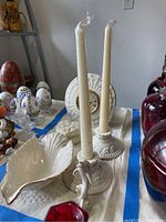 Photo showing white ceramic Lenox quartz clock, two candle sticks with candles, white shell shaped candy dish, and trinket box with floral design lid, also red glass vase partially visible