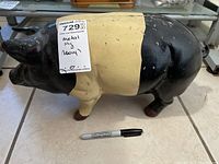 Side view of painted metal pig on tile floor with a Sharpie marker for scale