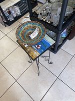 Side view of mosaic glass top table with metal base and decorative bowl on top, adjacent to shelving with glassware.