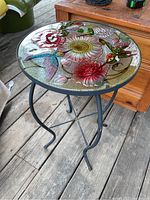 Full view of round glass topped patio table showing colorful hummingbird and floral design and metal base with curved legs.