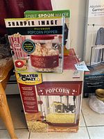Box of Sharper Image microwave popcorn popper with red lid and 6 paper popcorn bags included, placed on top of another box for popcorn machine.