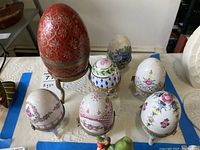 Overview of seven small porcelain eggs with floral and geometric patterns, plus one large red egg on metal toned stand.