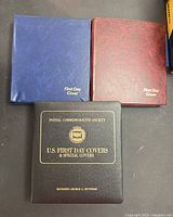 Three binders shown closed; black 'Postal Commemorative Society' binder with gold text, blue and red 'First Day Cover' binders.