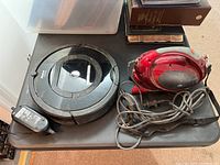 Photo showing iRobot Roomba 17062 robot vacuum and Durabrand red handheld vacuum with cords on a black surface.