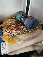 Folded textiles including vintage tapestry or blanket with fringe, packaged Brach towel and cream textiles, rolled Twister rug visible