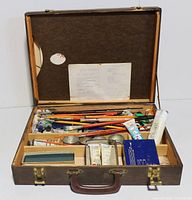 Open wooden case showing organized art supplies including paint brushes, paint tubes, and pencils inside compartments.