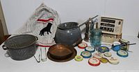 Photo showing large graniteware pot, gray metal basin, copper pan, glass jars, red handled utensil, vintage apron, and various collectible lids and tins.