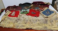 Overview photo of multiple vintage needlepoint and tapestry pieces spread out showing Scottie dogs, floral, and large classical scene on tan cloth.