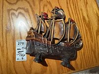 Photo of a cast iron doorstop shaped like an old sailing ship with three sails, the tips painted red, resting on a wooden floor.