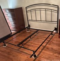 Metal bed frame with arched metal headboard positioned against a wall. Room background includes hardwood floor, chest of drawers, and nightstand.