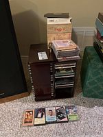Photo of dark wood spinning CD stand loaded with CDs and five cassettes laid out on carpet.