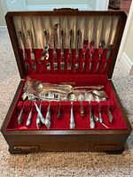 Open wood box showing satin-lined interior containing flatware set including forks, knives, tablespoons, teaspoons, and serving utensils arranged neatly inside.