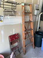 Photo showing a wood ladder standing upright with visible paint marks and a red metal-frame two-step stool leaning against the wall; both items are in a garage environment.