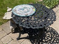 Front angle of ornate black cast metal garden table with mosaic hummingbird plaque and beige Chinese-style garden lantern visible beside it on stone patio