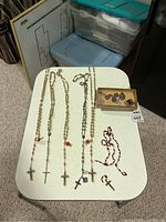 Four rosaries with cross pendants and one broken red rosary on white table, small box with religious pendants beside them.