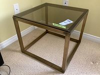 Front angle showing brass coloured metal frame base and grey glass top, resting on carpeted floor near outlet.