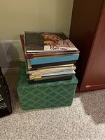 Stack of mixed vinyl records on top of a green patterned storage ottoman.