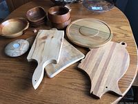 Photo showing an assortment of wooden cutting boards, wooden and ceramic bowls, a wooden lazy Susan, and a stone candle holder on a wooden table.