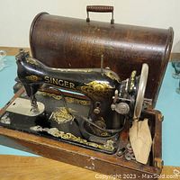 Black Singer sewing machine with gold details sitting on wooden base, with partial view of wooden carrying case behind it