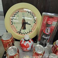 Photo showing Coca-Cola wall clock, collectible cans, soccer ball bottle, bottle opener in package, and keychain on display.