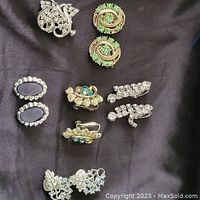 Top view of multiple pairs of vintage clip on earrings on black fabric, showing various designs with green stones, dark cabochons, and clear rhinestones.