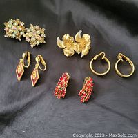 Full view of the five pairs of clip-on earrings arranged on black fabric background, showing floral, hoop, red stone cluster, and geometric enamel styles.