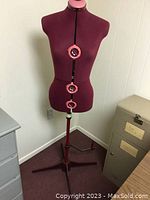 Front view of vintage burgundy fabric dress form with adjustable dials and tripod base near filing cabinet and desk in a room.