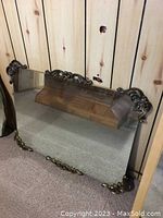 Front view showing large rectangular mirror with ornate metal frame, intricate scroll and floral design at top and bottom, resting against wood panel wall.