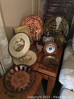 Photo showing a variety of brass and copper decorative plates, a lamp with brass base, a small barometer or clock, and a twisted edge tray placed on a wooden table next to a couch.