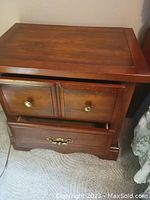 Nightstand with three drawers including two smaller upper drawers and one larger lower drawer, wood finish with brass and metal handles, shows some use marks and wear.