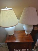 Pair of white glass lamps with pleated fabric shades on a wooden nightstand, one lamp illuminated to show working condition.