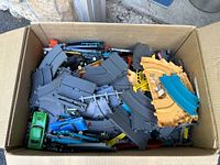 Box containing numerous plastic toy race track pieces in gray, blue and yellow colors, mostly curved tracks.