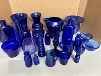 Wide view showing 26 blue glass and ceramic pieces of various shapes and sizes including vases, bottles, jars, cups, and pitchers all in shades of cobalt blue.