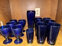 Photo showing all blue glassware including 6 pint glasses, 6 goblets, 1 tall vase, and 1 juice glass arranged on wooden shelves