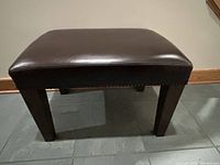 Front view showing square brown leather ottoman with wooden legs and nailhead trim detail