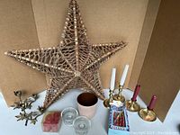 Photo showing large wicker star, various candleholders in bronze and brass, small ceramic pot, glass votive holders, boxes of candles and cranberry taper candles.