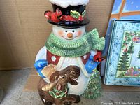 Ceramic snowman cookie jar with black top hat, green scarf, red cardinals, brown dog, and small Christmas tree