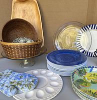 Photo showing assortment of servingware including wicker basket on clear glass pedestal, wood platter, clear glass plates, blue ceramic plates, deviled egg platter, and colorful tropical plates.