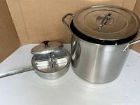 Large stainless steel pasta pot with lid and small divided pot with lid placed side by side against cardboard background.