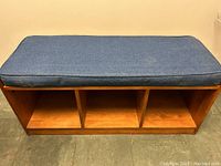 Front view of wooden shoe bench with denim-colored cotton cushion on top, showing three open cubbies for shoe storage.