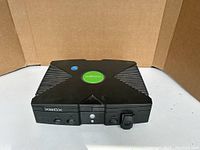 Front perspective of the black original Xbox console showing the 'X' top design and green Xbox logo in the center.