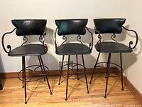 Three wrought iron bar stools with black leather padded seats and backs against a wall on a wooden floor, showing front and side details including scroll armrests and tapered legs with footrests.