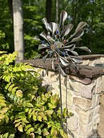 Large kinetic moving metal garden sculpture with curved blades and embedded colorful glass beads, 6 feet tall and 2 feet wide, shown outdoors against stone wall and greenery.