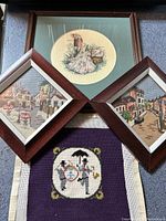 Framed needlepoint artworks featuring three pictures: one circular floral needlepoint with basket, two diamond-shaped urban needlepoints, and one of two dancing characters on purple fabric.