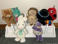 Photo of the handcrafted white wooden puppet rack displaying seven assorted puppets including bear, lion, green dinosaur, seal, Clifford the Big Red Dog, purple rhino, and white rabbit puppets.