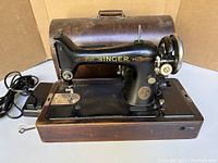 Front view of Singer sewing machine mounted on wooden base with cover behind it, showing pedal cord and visible handle on cover.