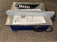 Tile saw unit with power cord and handle, aluminum guide rail placed on top, and instruction manual visible.