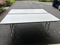Two white folding tables with sturdy metal legs shown outdoors on asphalt surface, viewed from back and side angles.