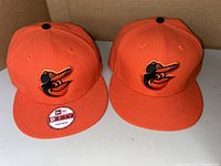 Two orange Baltimore Orioles caps side by side on a white surface, showing embroidered logo and snapback style. One cap has a 9FIFTY sticker on the brim.